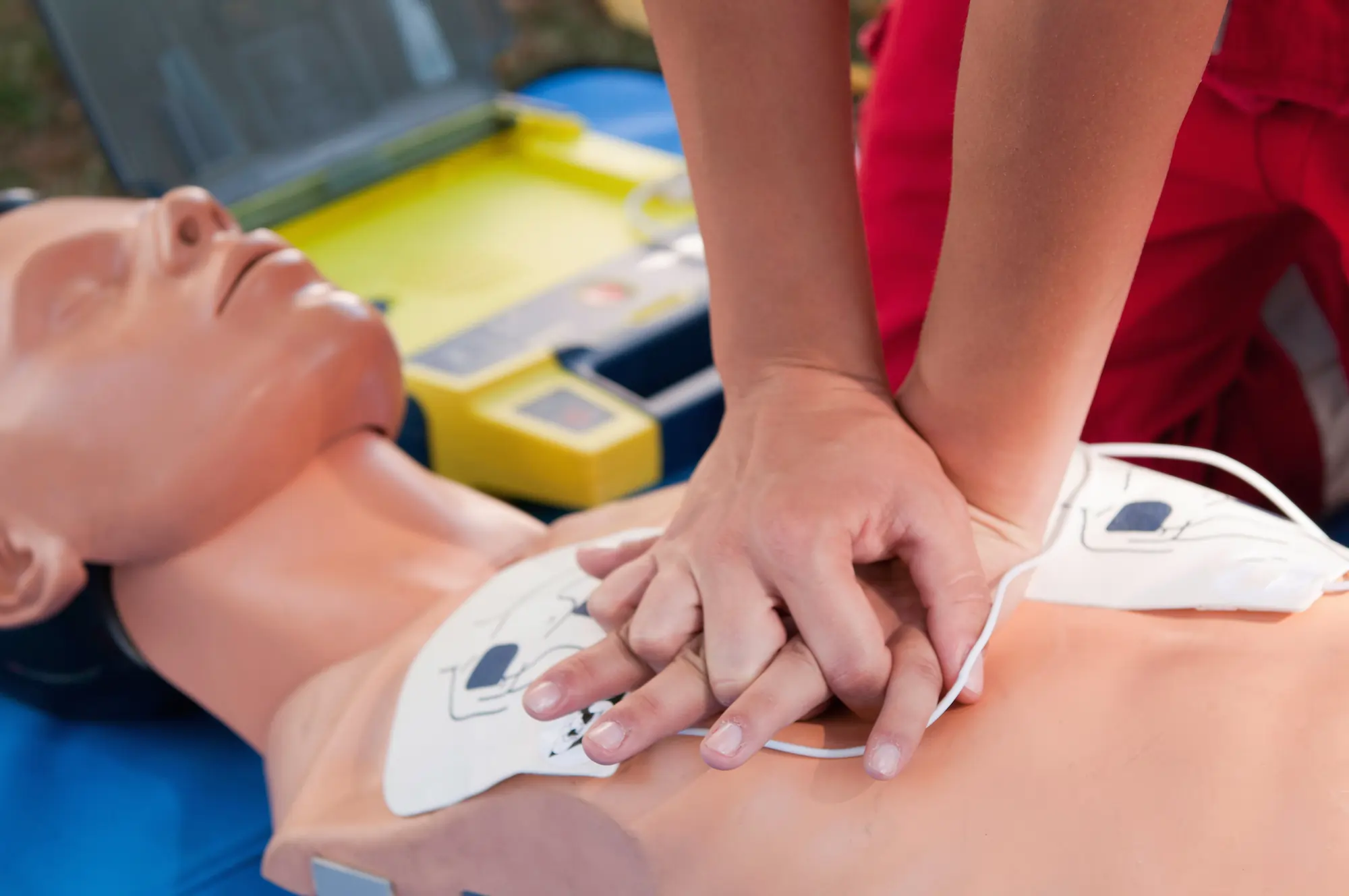 A person practises CPR on a medical manikin with a defibrillator nearby at Mobile First Aid CPR Peregian Springs, Sunshine Coast; hands are positioned on the manikin’s chest, pressing down for chest compressions.