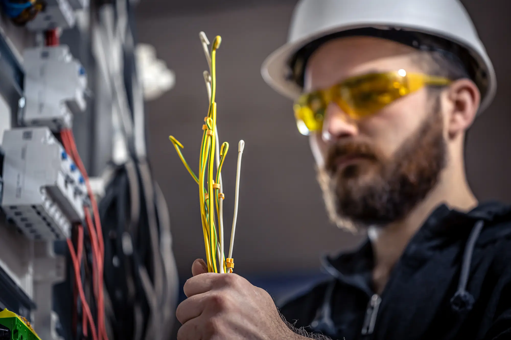 A man wearing a white hard hat and yellow safety glasses holds several electrical wires whilst working in front of an electrical panel, demonstrating skills from the Work Safely Near Live Electrical Equipment course in Peregian Springs, Sunshine Coast.