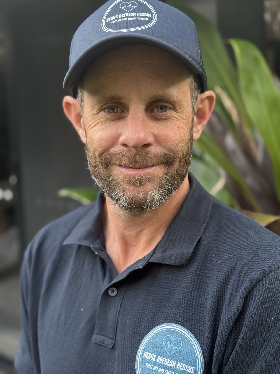 A man wearing a navy shirt and cap with Resus Refresh Rescue stands outdoors, smiling slightly, likely participating in first aid training or a CPR course, with greenery and a building behind him.