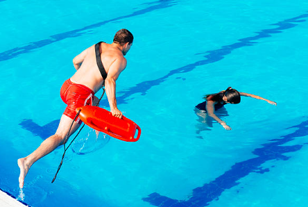 A lifeguard trained in CPR and First Aid Training Peregian Springs jumps into a swimming pool, holding a rescue buoy, heading towards a child who appears to be struggling in the water.