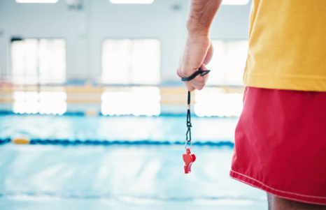 A person in a yellow shirt and red shorts stands poolside, holding a red whistle on a black lanyard, suggesting they are a lifeguard with CPR and First Aid Training in Peregian Springs, overseeing an indoor swimming pool.