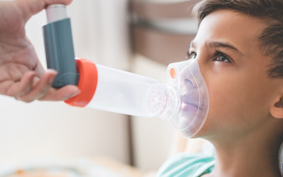 A young boy wearing an inhalation mask receives asthma medication from an inhaler held by an adult hand—an important scenario often covered in CPR and First Aid Training Peregian Springs. The boy looks up as he breathes through the mask.