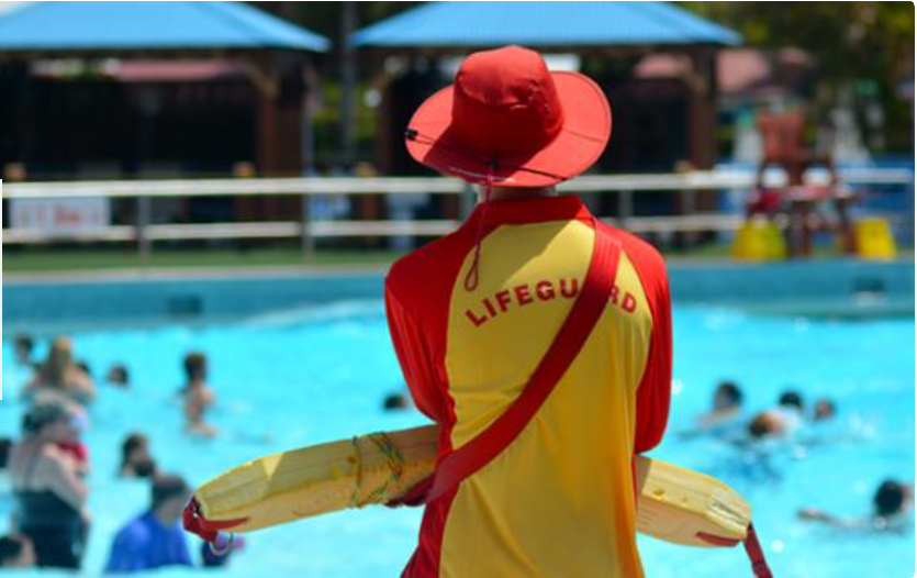 A lifeguard in a red and yellow uniform and hat stands by a busy swimming pool, watching over swimmers on a sunny day, demonstrating the vigilance learnt through CPR and First Aid Training Peregian Springs.